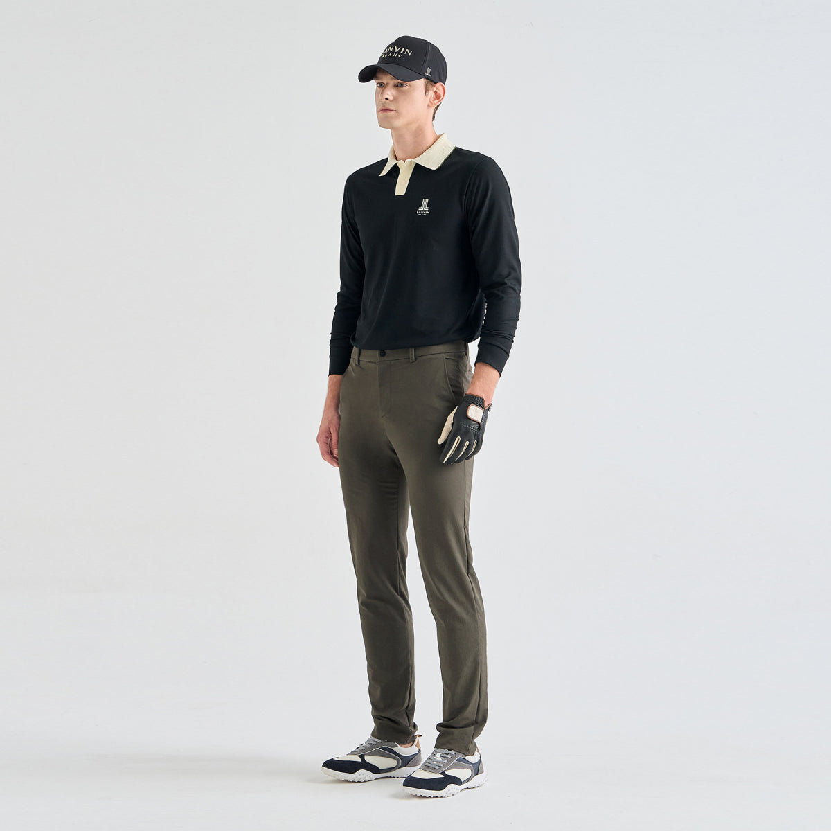 A male model stands in a studio wearing a black long-sleeve polo shirt with a light beige collar, olive green pants, a black cap, a black and white golf glove, and black and white sneakers.
