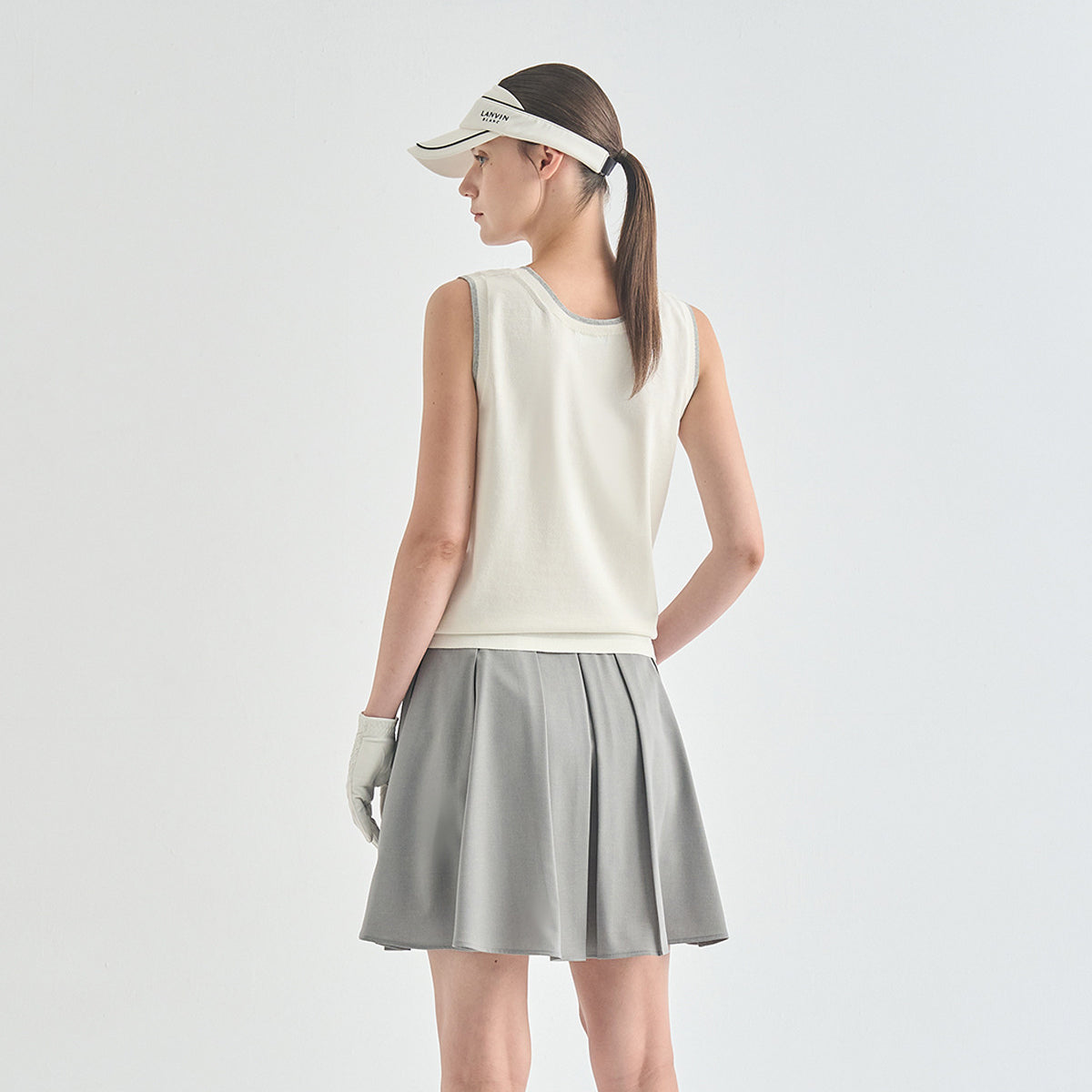 A woman modeling a sleeveless off-white top and pleated gray skirt set, wearing a white visor and light gray glove, photographed against a clean light gray studio background.