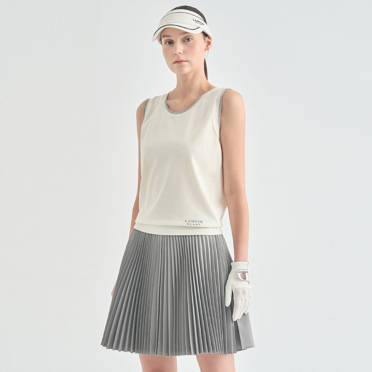 A model wearing a sleeveless off-white athletic tank top, a gray pleated tennis skirt, a white visor, and a white golf glove, standing against a light gray studio background.