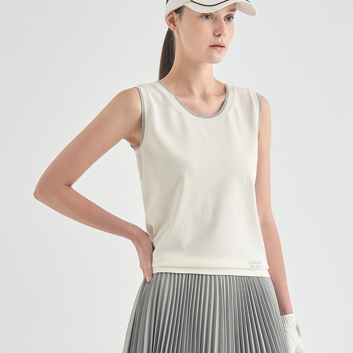 A woman modeling a sleeveless athletic tank top in off-white with gray trim, paired with a gray pleated skirt and a white visor, against a clean light gray studio background.