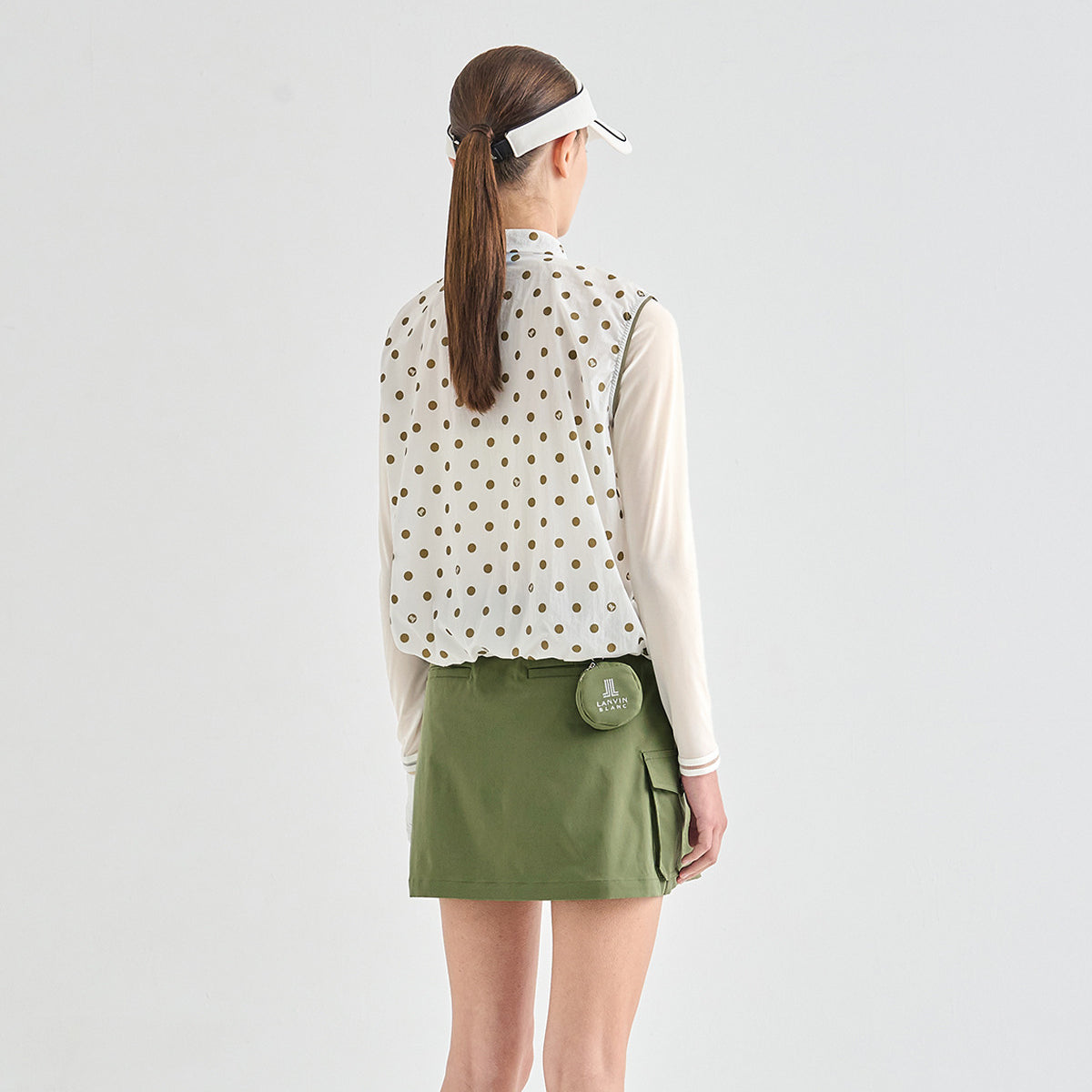 A woman is shown from behind wearing a sleeveless white vest with a gold polka dot pattern over a cream long-sleeve shirt, paired with an olive green skort. She is wearing a white visor and standing against a solid light gray background.