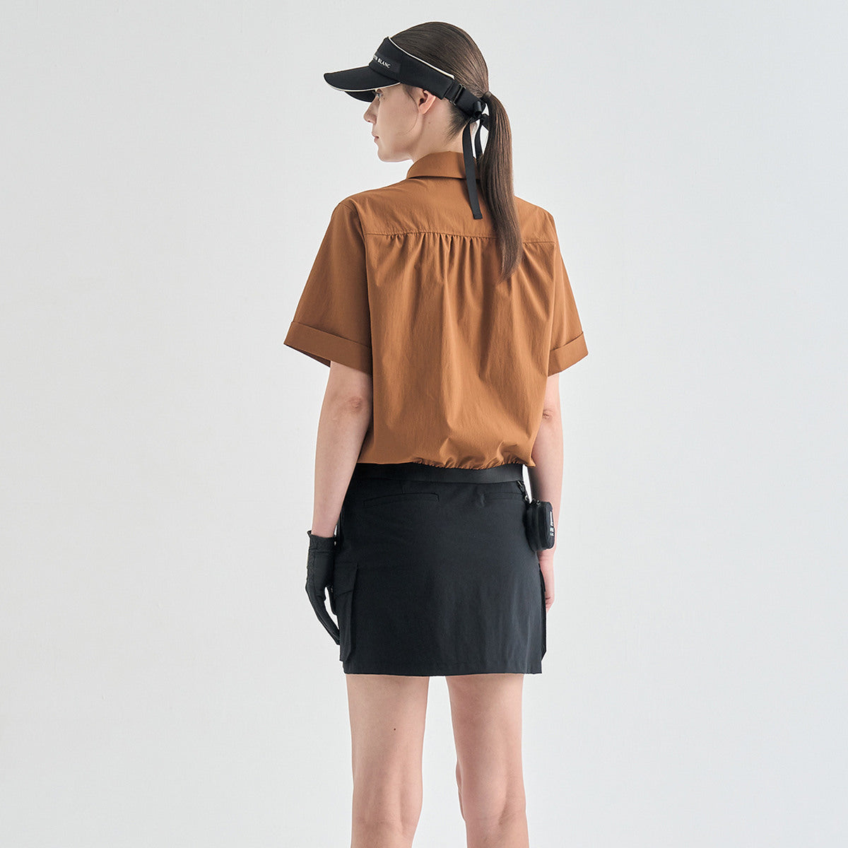 A woman shown from the back wearing a brown short-sleeve collared shirt, a black mini skirt, black gloves, a black visor, and a black ponytail, standing against a light gray studio background.