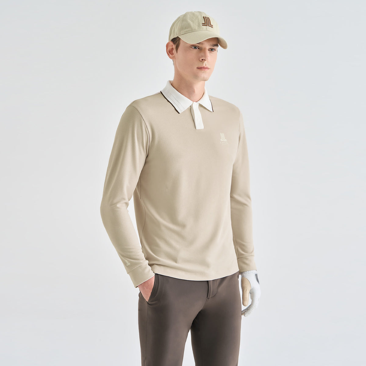 A man modeling a beige long-sleeve polo shirt with a contrasting white collar, paired with brown pants, a beige cap, and a white golf glove, against a light gray studio background.