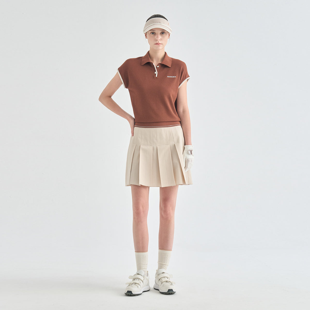 A model stands against a light gray studio background wearing a brown short-sleeve polo shirt, a beige pleated tennis skirt, a beige visor, white gloves, white socks, and white sneakers.
