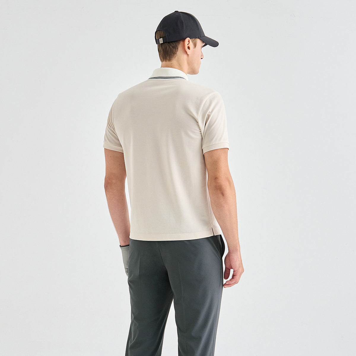 A man shown from the back wearing a light beige short-sleeve polo shirt, dark gray trousers, and a black baseball cap against a plain light gray studio background.