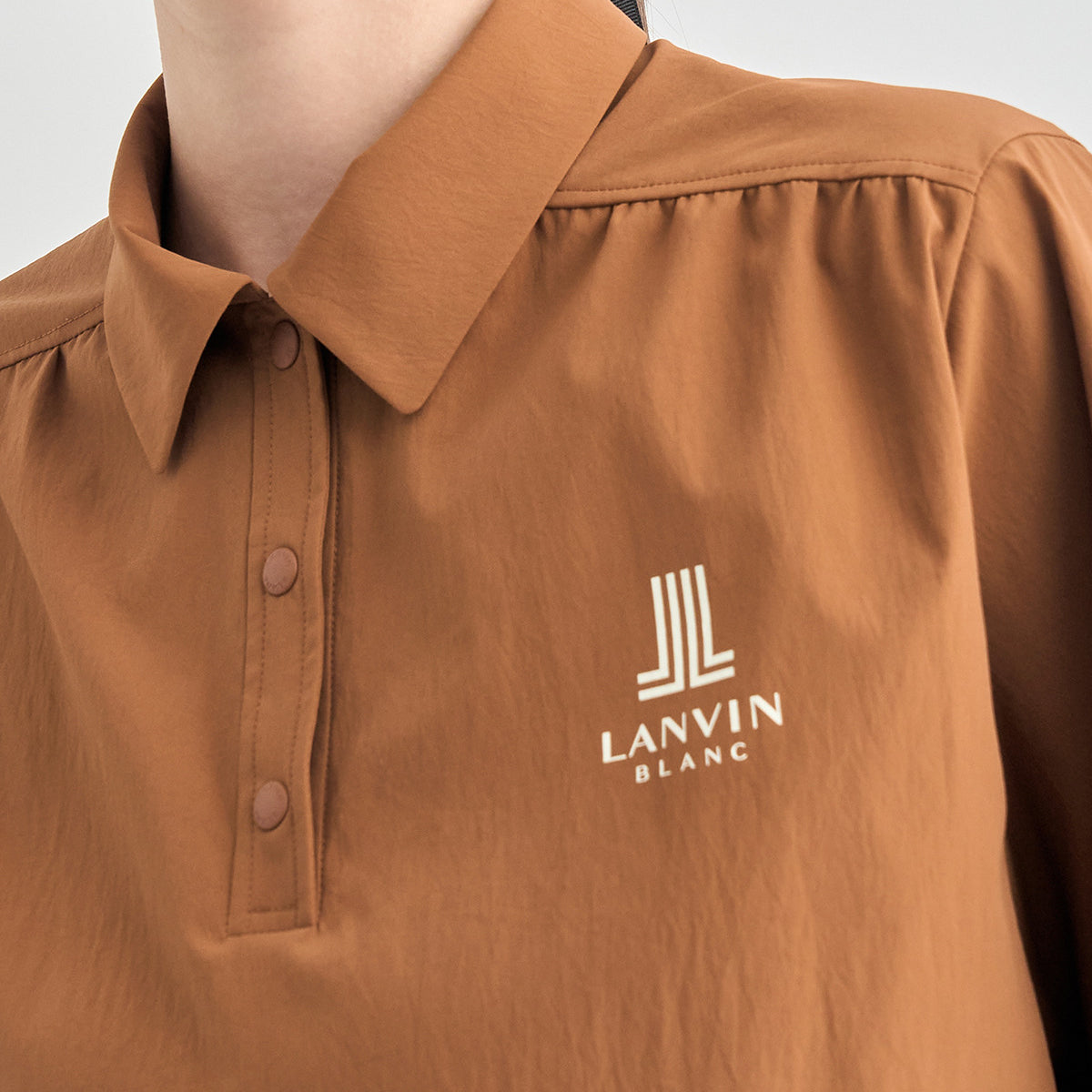 Close-up of a person wearing a tan-brown collared button-up shirt with a white embroidered Lanvin Blanc logo on the chest.