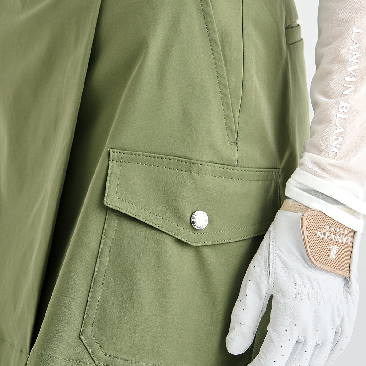 Close-up of an olive green jacket with a flap pocket and silver snap, shown next to a white golf glove with Lanvin Blanc branding.