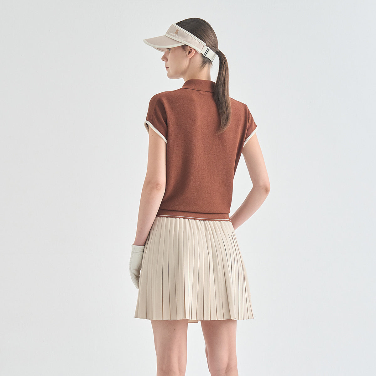 A woman shown from the back wearing a fitted short-sleeve brown top with cream trim, a pleated cream skirt, a cream visor, and cream gloves against a light gray studio background.