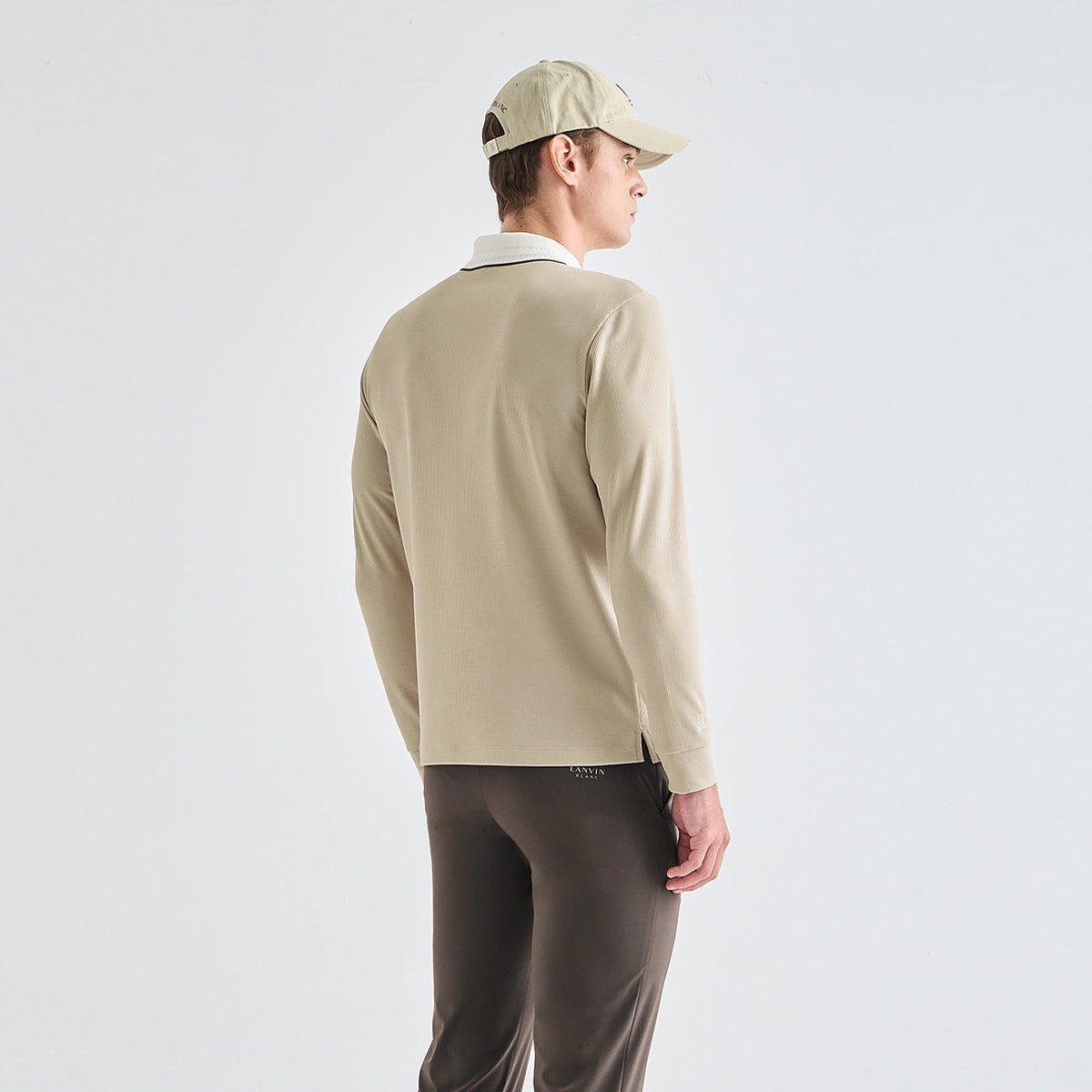 A man shown from the back wearing a light beige long-sleeve polo shirt, dark brown pants, and a beige baseball cap, against a plain light gray studio background.