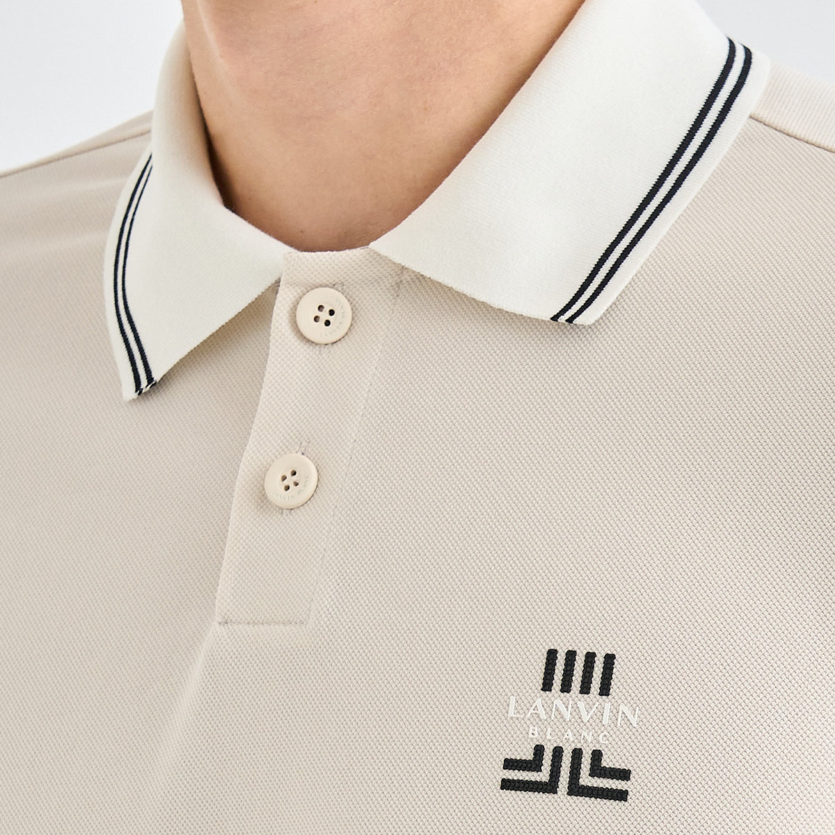 Close-up of a person wearing a light beige polo shirt with a white collar trimmed in black double stripes. The brand logo 'LANVIN BLANC' is embroidered in black on the chest.