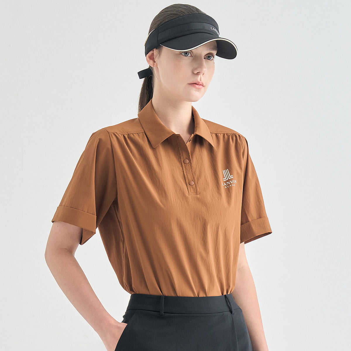 A woman wearing a short-sleeve brown polo shirt with a small white embroidered logo on the chest, paired with black pants and a black visor, photographed against a light gray studio background.