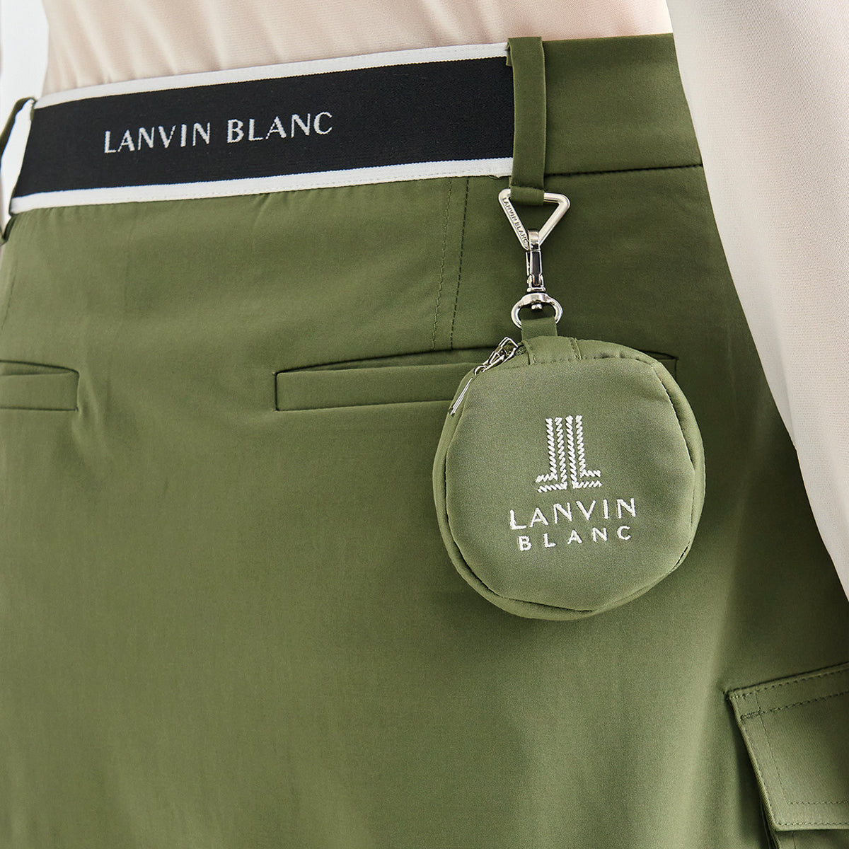Close-up of olive green shorts with a black waistband labeled LANVIN BLANC and a matching small round pouch clipped to the side pocket, also branded LANVIN BLANC.