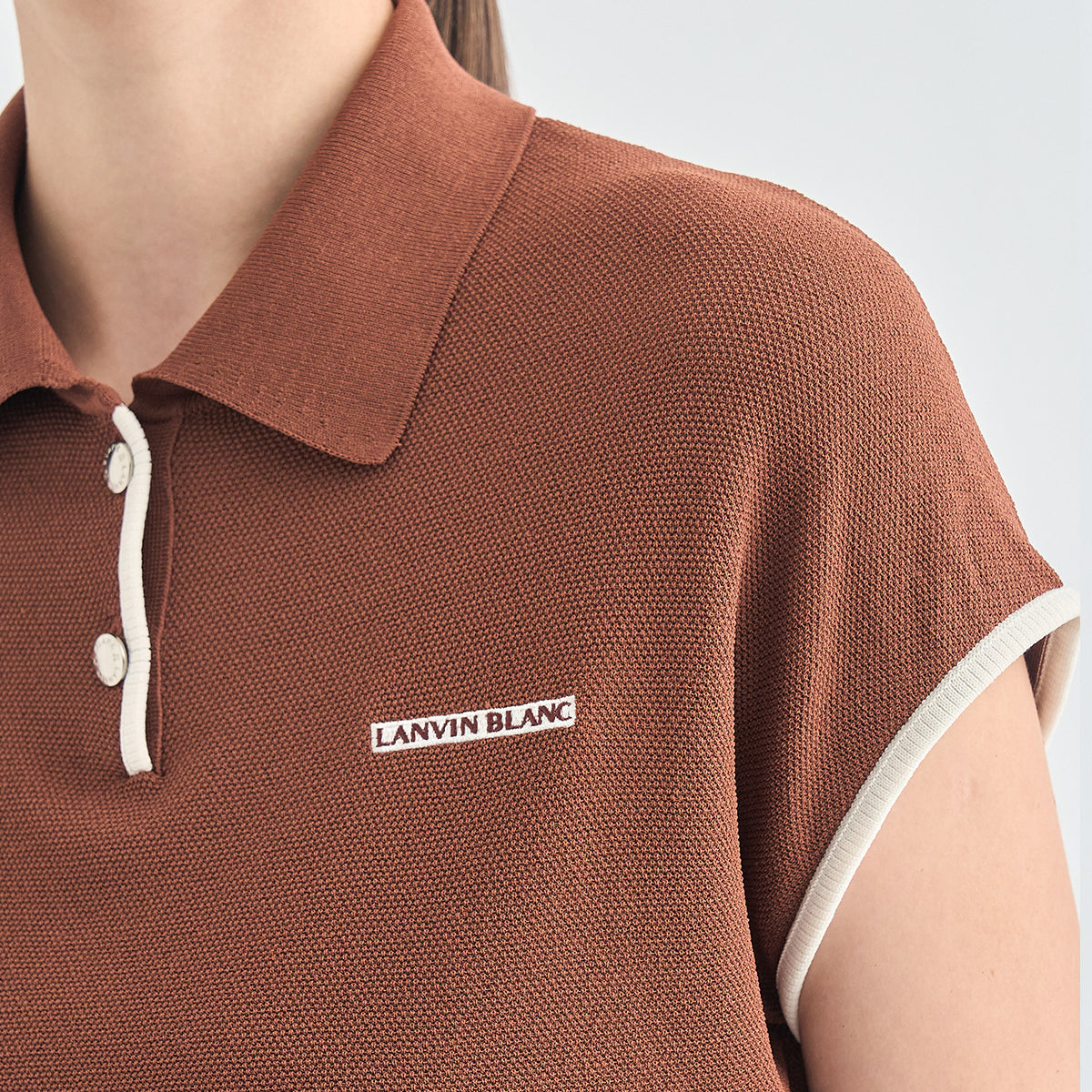Close-up of a person wearing a brown short-sleeve polo shirt with white trim on the collar and sleeve cuffs, featuring a white embroidered 'LANVIN BLANC' logo on the chest.