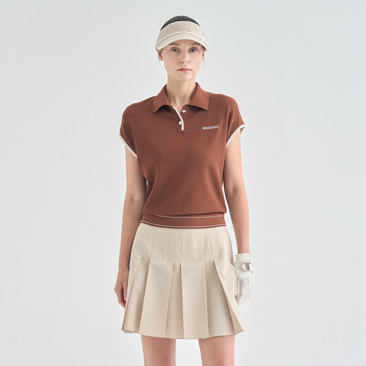 A model stands against a light gray studio background wearing a short-sleeve brown knit polo shirt with a small white logo on the chest, a pleated beige tennis skirt, a beige visor, and a white golf glove.