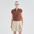 A model stands against a light gray studio background wearing a short-sleeve brown knit polo shirt with a small white logo on the chest, a pleated beige tennis skirt, a beige visor, and a white golf glove.