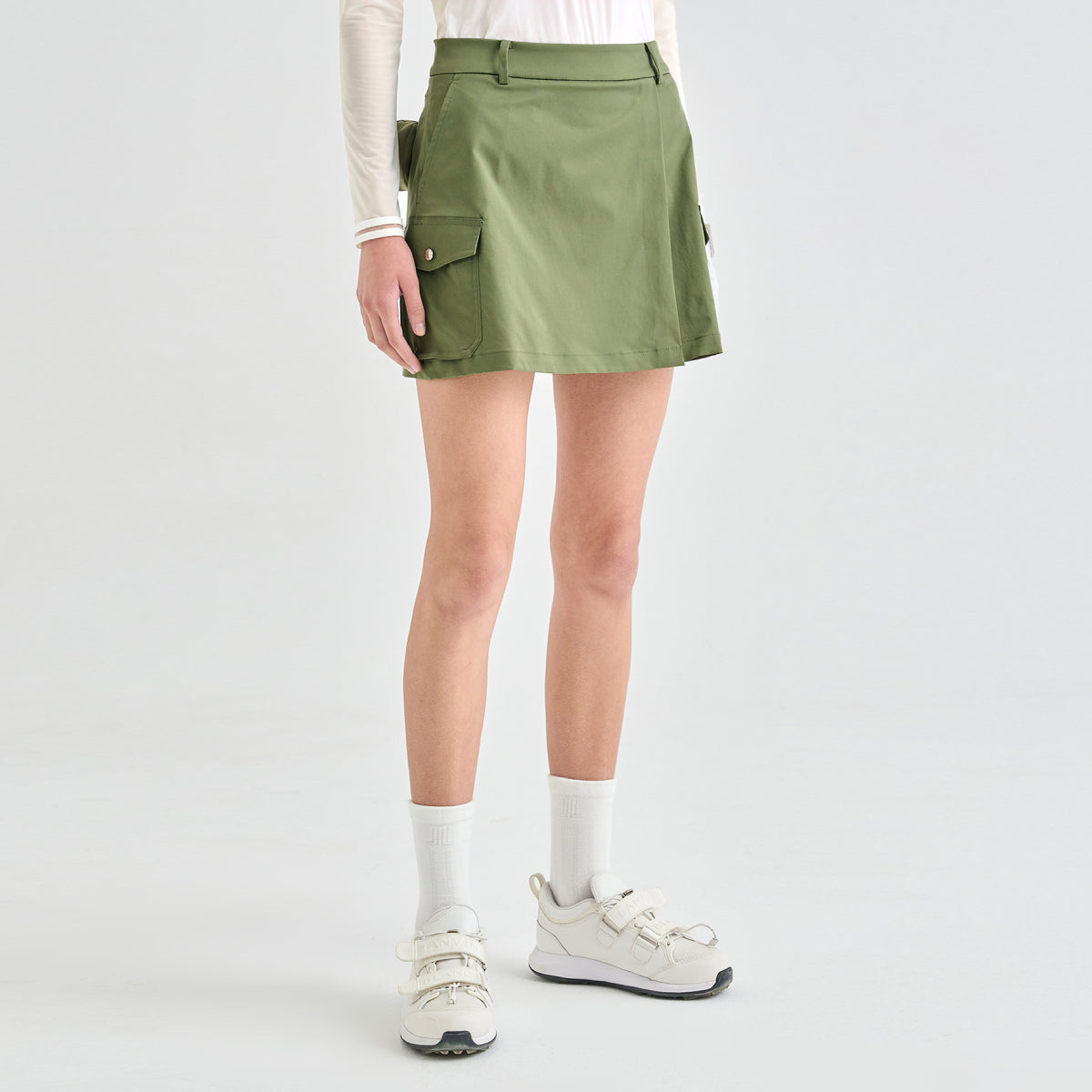 A model wearing an olive green cargo-style mini skirt with flap pockets, paired with white socks and white sneakers, shown from the waist down on a light gray studio background.