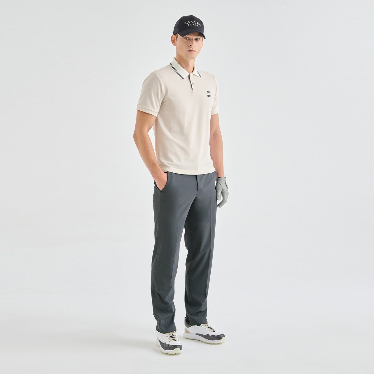 A man modeling a light beige polo shirt, dark gray trousers, a black cap, and a gray golf glove against a plain light gray studio background.