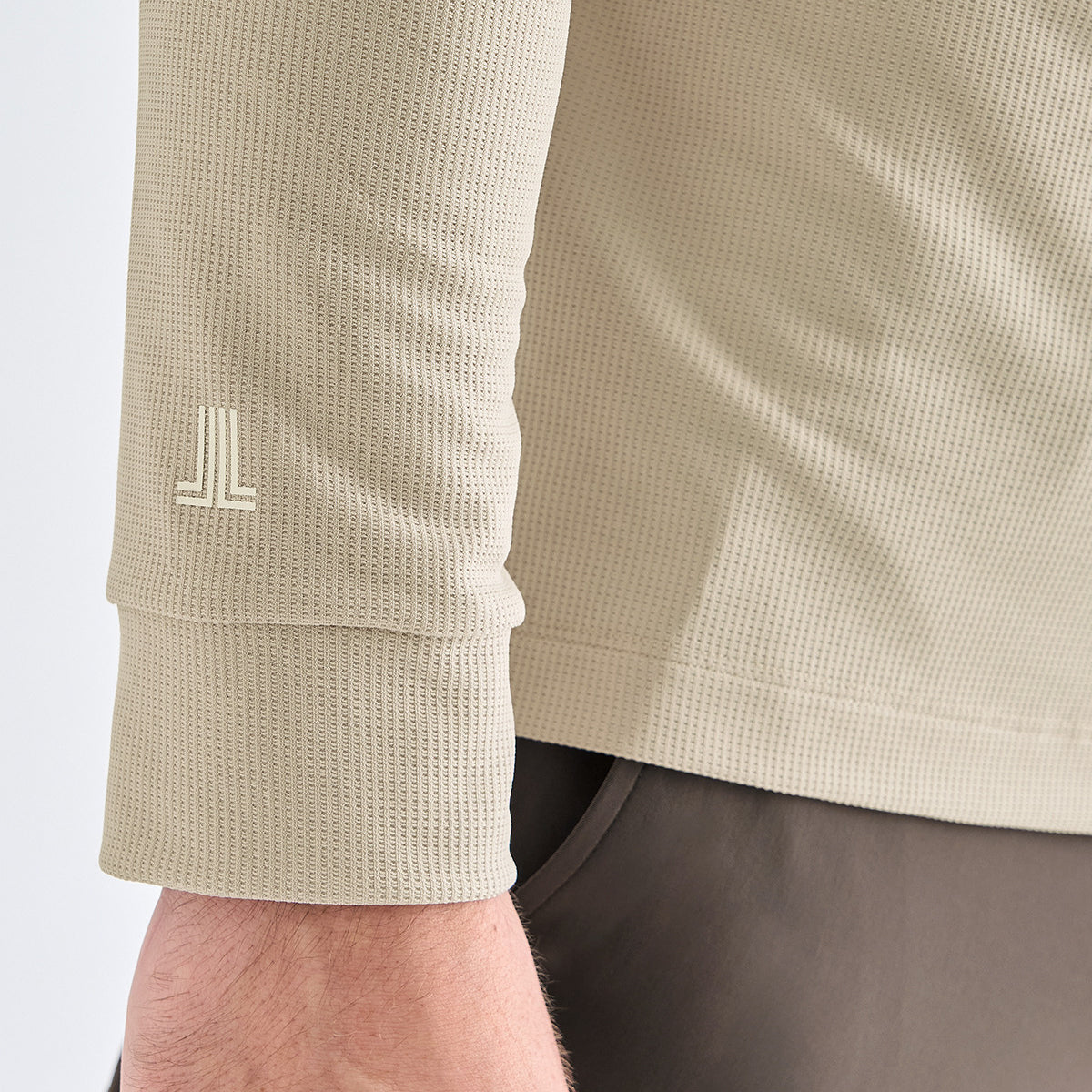 Close-up of a beige waffle-knit long-sleeve top worn by a model, showing the textured fabric and a small white stylized logo on the cuff.