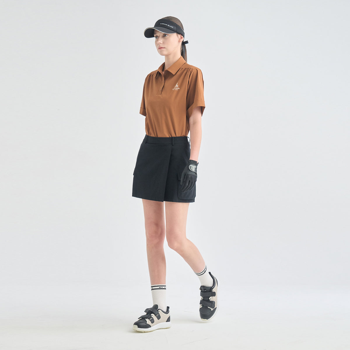 A model wearing a brown short-sleeve polo shirt, black athletic shorts, a black visor, a black golf glove, and black-and-white sneakers, standing against a plain light gray studio background.