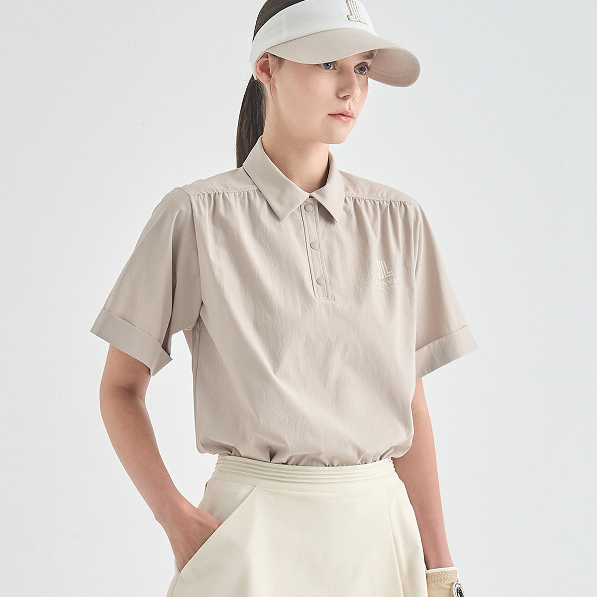 A woman wearing a light beige short-sleeve polo shirt with a small tonal embroidered logo on the chest, paired with a matching light beige skirt and a white visor, set against a plain light gray studio background.