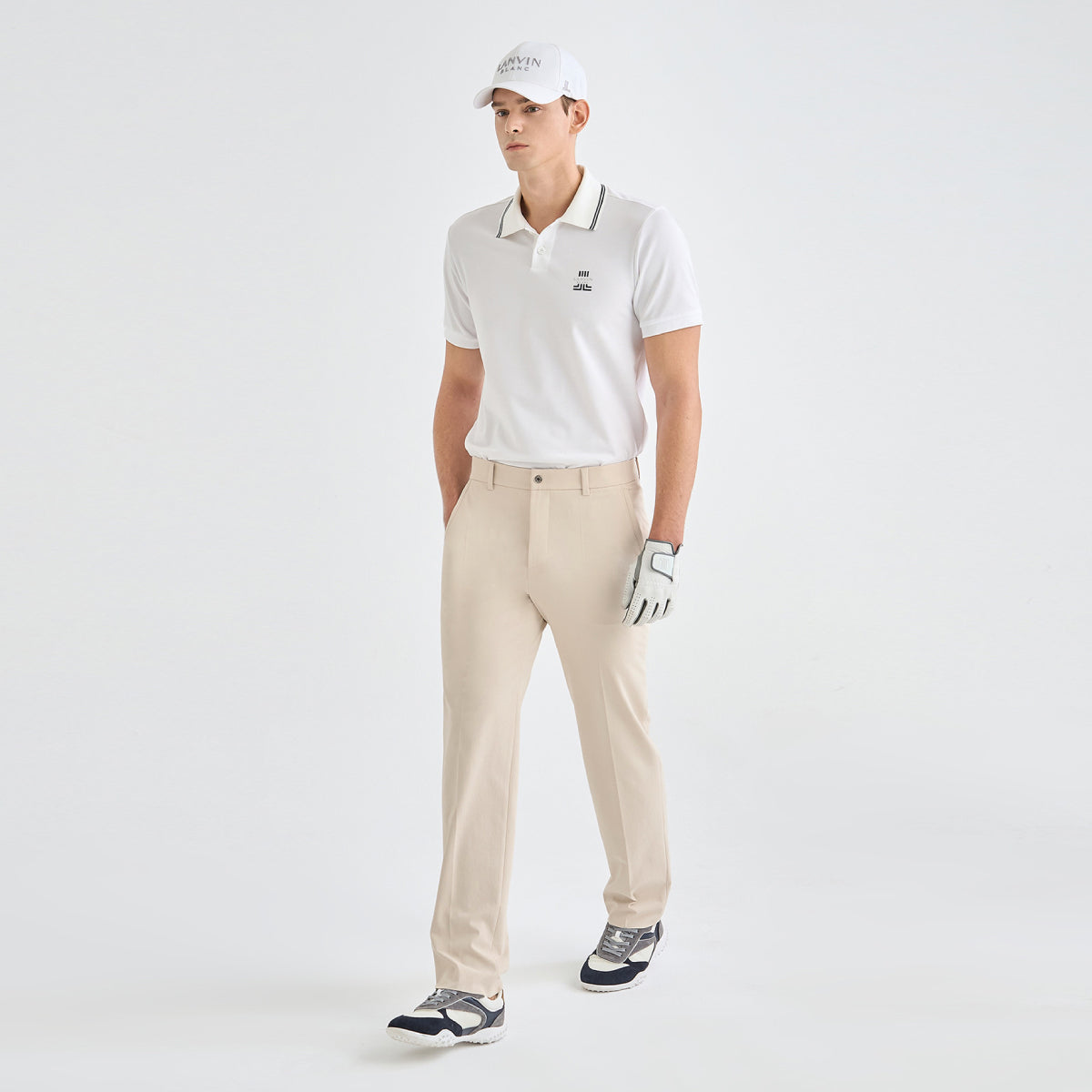 A man modeling a white short-sleeve polo shirt with dark piping, beige golf pants, a white cap, and a golf glove against a light gray studio background.