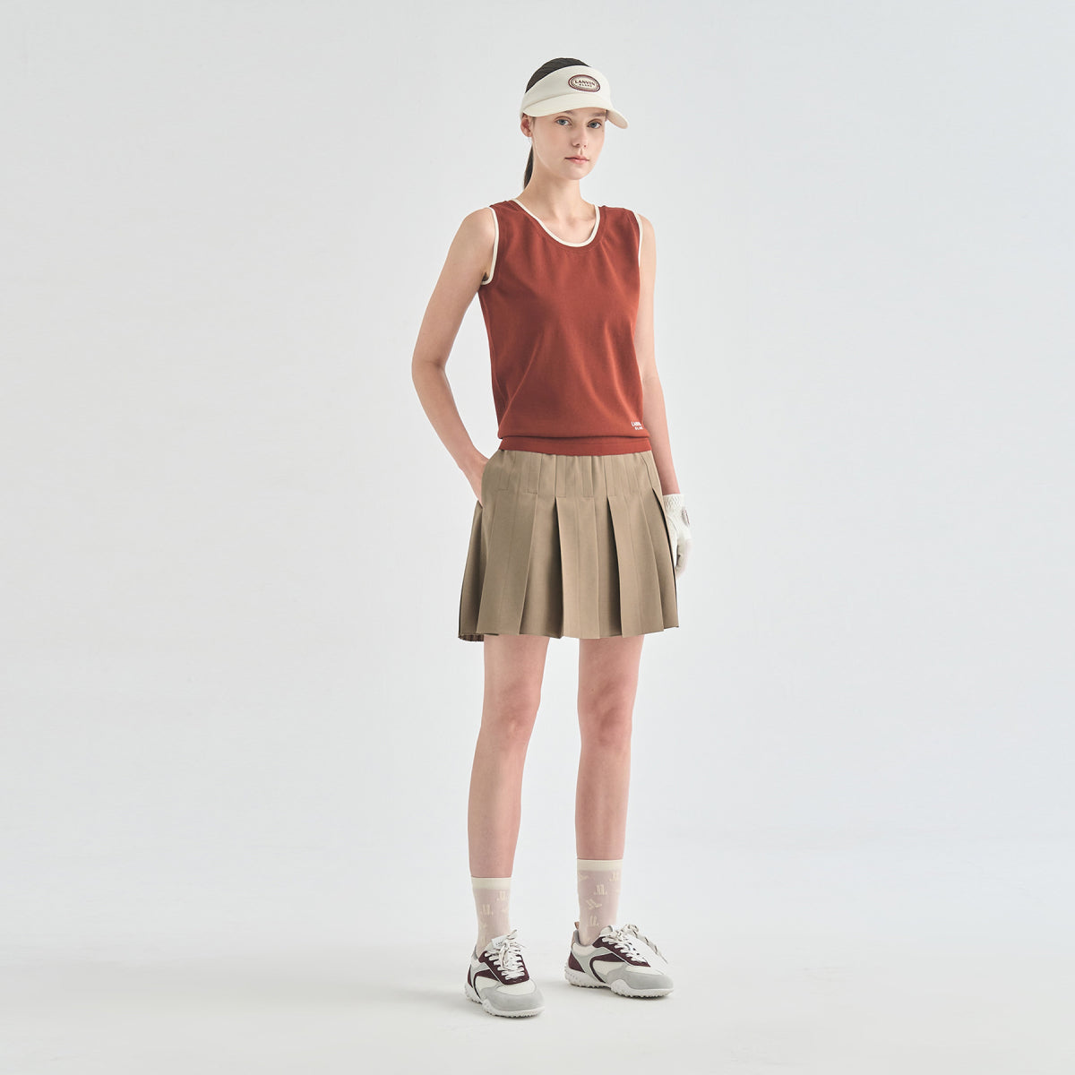 A model stands against a light gray studio background wearing a sleeveless rust-orange athletic top, a beige pleated tennis skirt, a beige visor, white socks, and white sneakers.