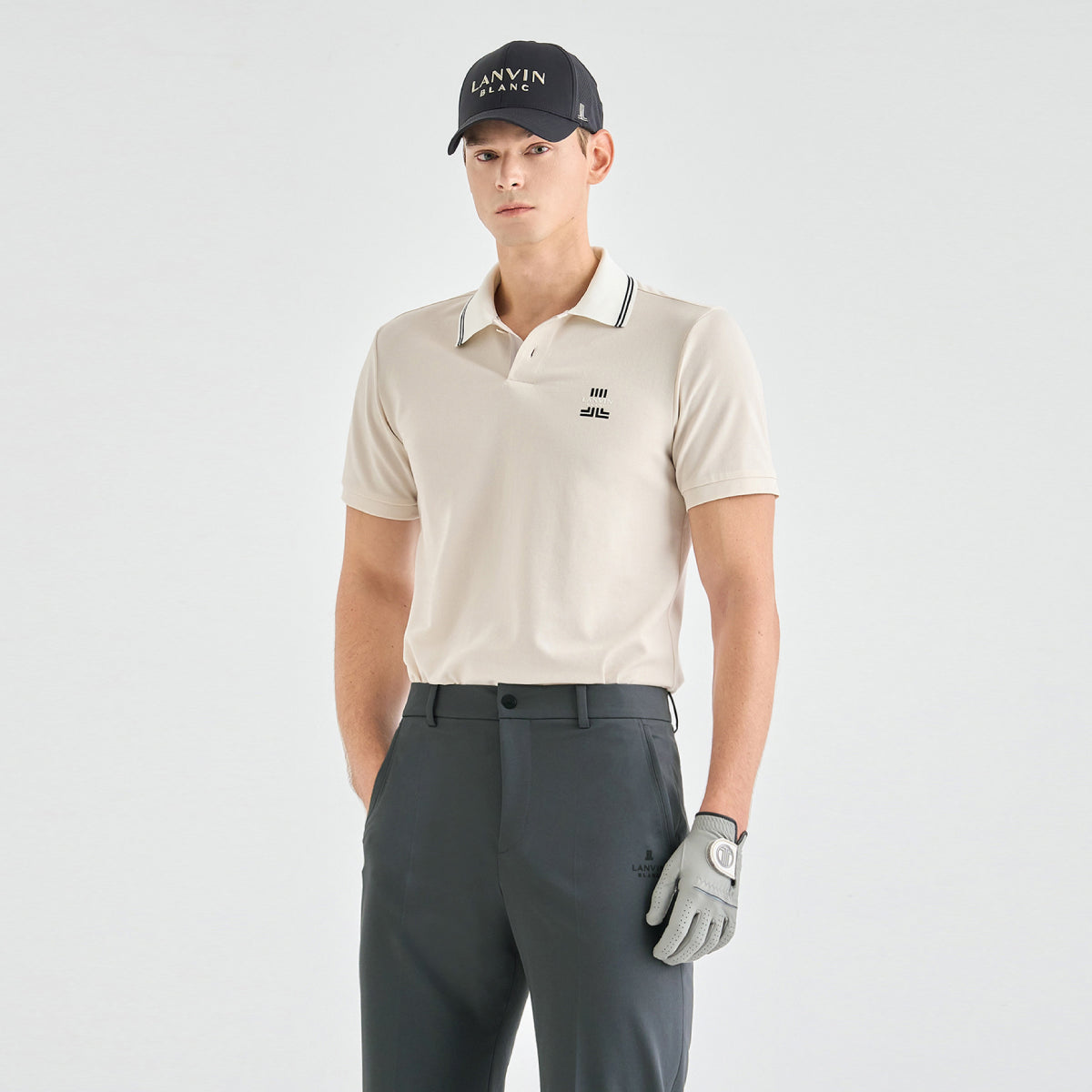 A male model wearing a light beige short-sleeve polo shirt with a dark green and white striped collar, paired with dark gray trousers, a black cap with 'LANVIN BLANC' text, and a light gray golf glove, against a plain light gray studio background.