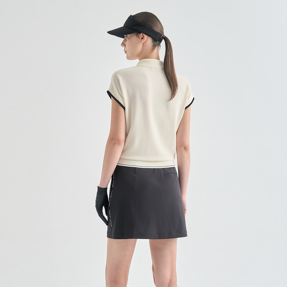 A woman shown from the back wearing a cream sleeveless knit polo-style top with black trim, a dark gray athletic skirt, black gloves, and a black visor against a light gray studio background.