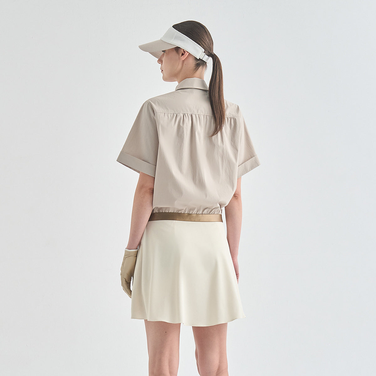A woman shown from the back wearing a beige short-sleeve collared shirt tucked into a cream tennis skirt, with a beige visor and matching glove, against a plain light gray studio background.