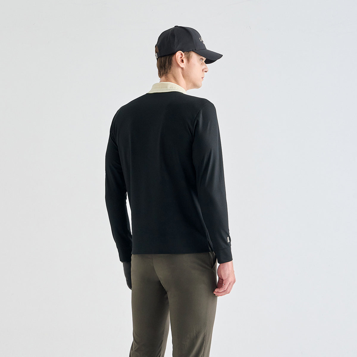 A man shown from the back wearing a black long-sleeve knit top with a light beige collar, olive green pants, and a black baseball cap, against a plain light gray studio background.