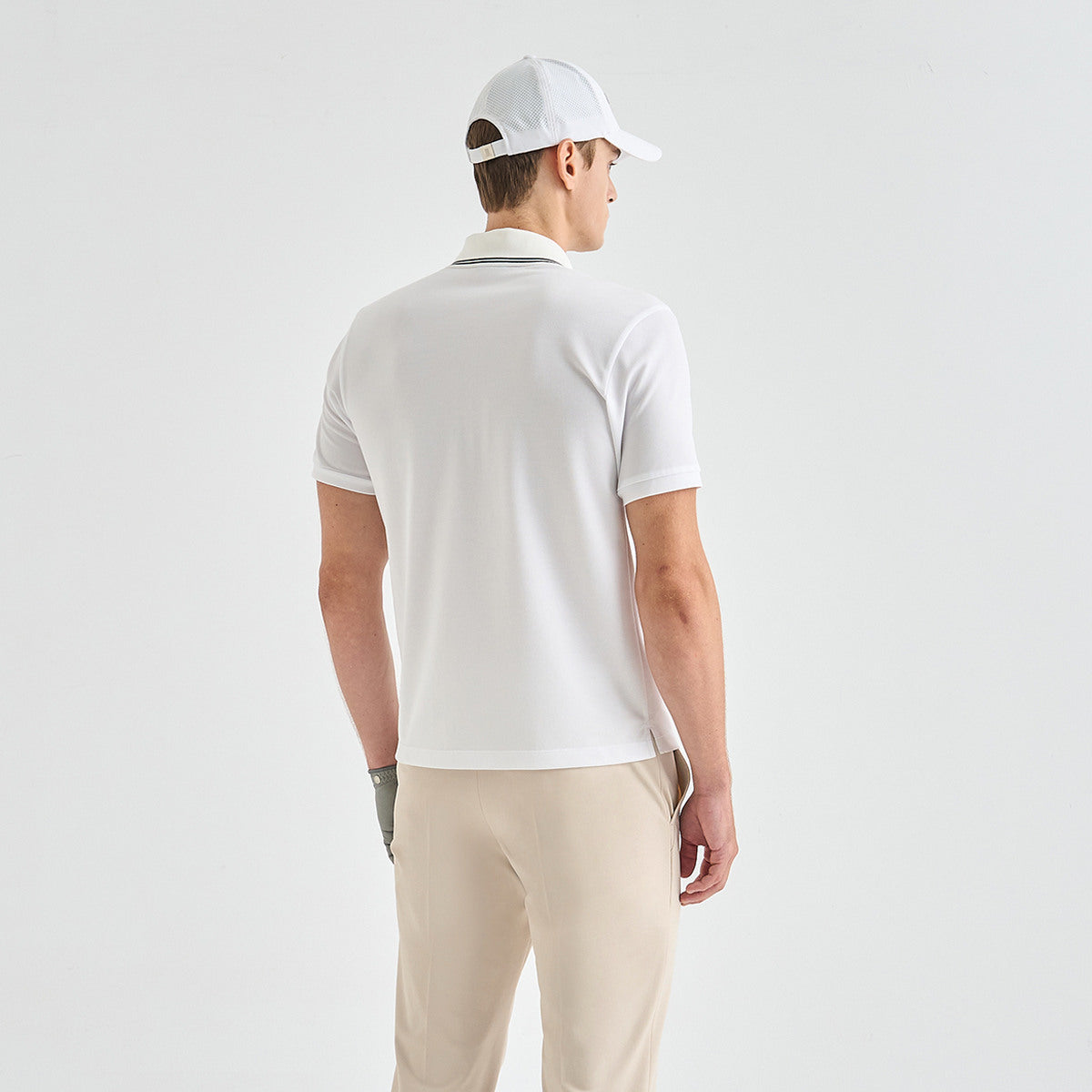A man shown from the back wearing a white short-sleeve polo shirt, beige trousers, a white baseball cap, and a gray golf glove, against a plain light gray studio background.