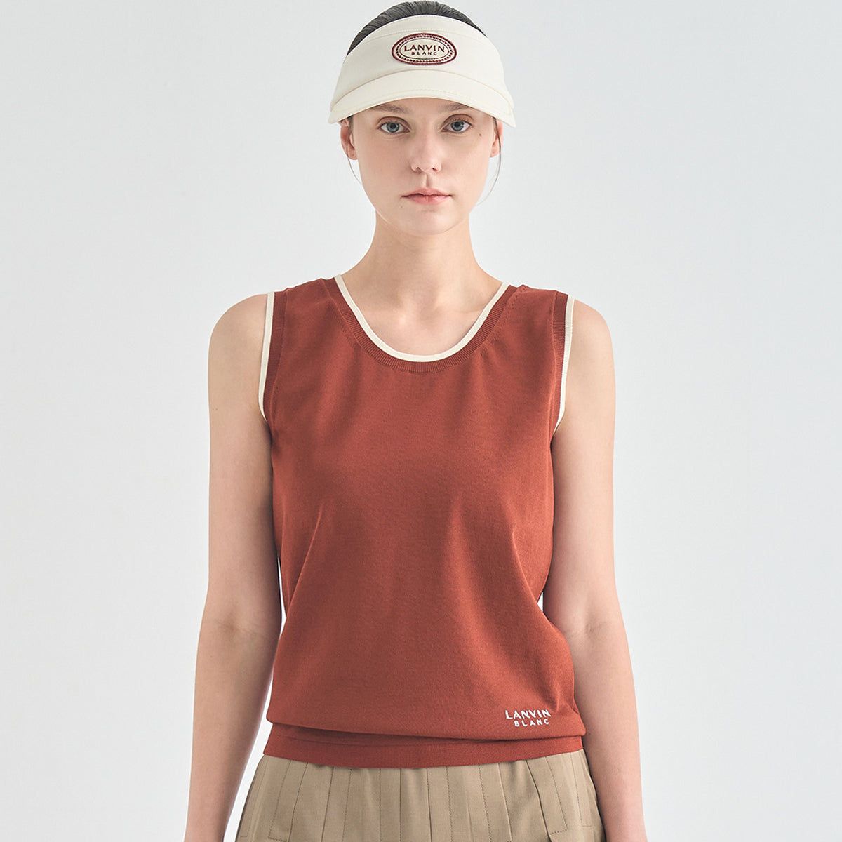 A model wearing a sleeveless rust-orange athletic tank top with cream trim, a cream visor with a visible Smith &amp; Wesson logo, and khaki pleated bottoms, shot against a light gray studio background.