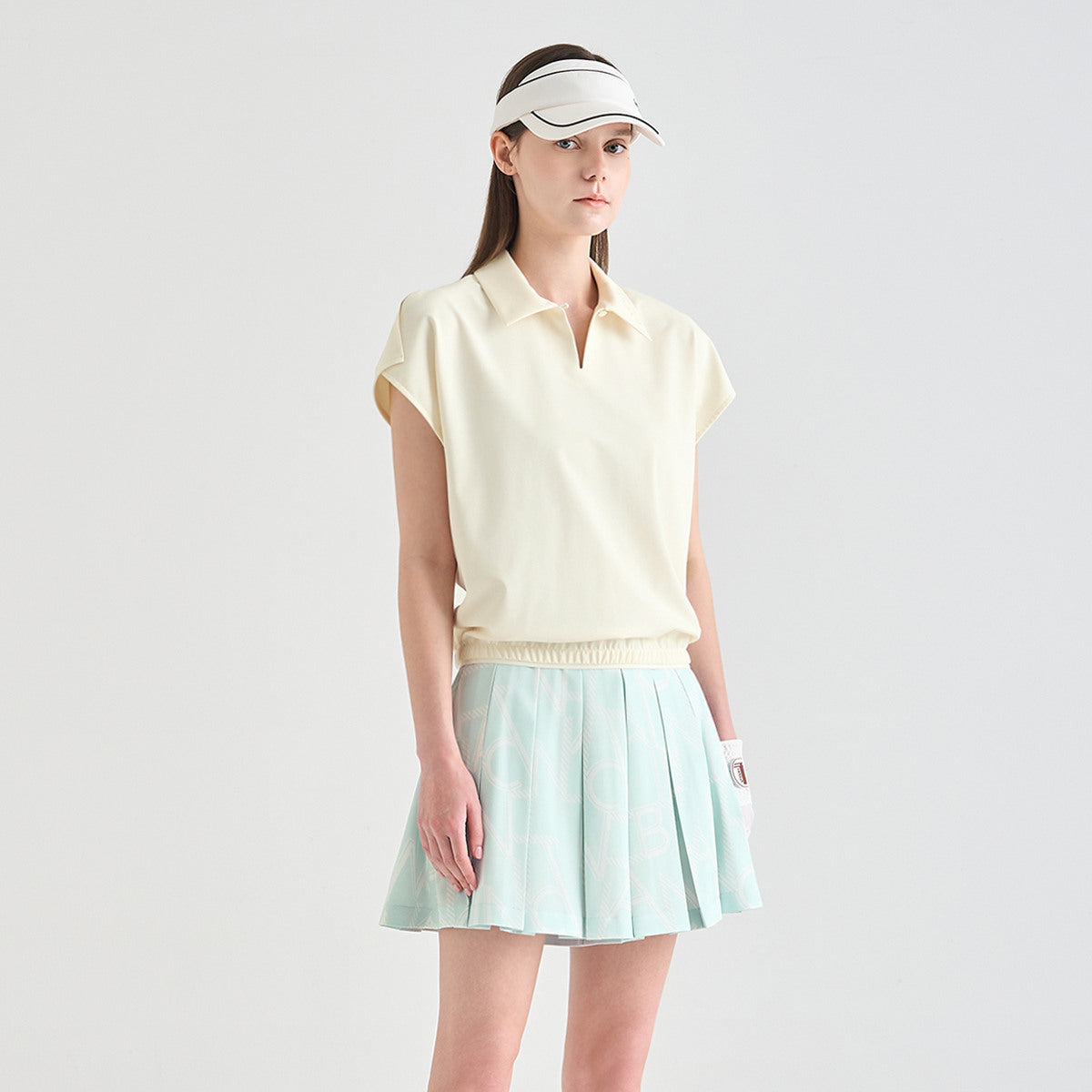 A woman modeling a light cream short-sleeve polo shirt and a light mint green pleated tennis skirt against a plain light gray studio background.