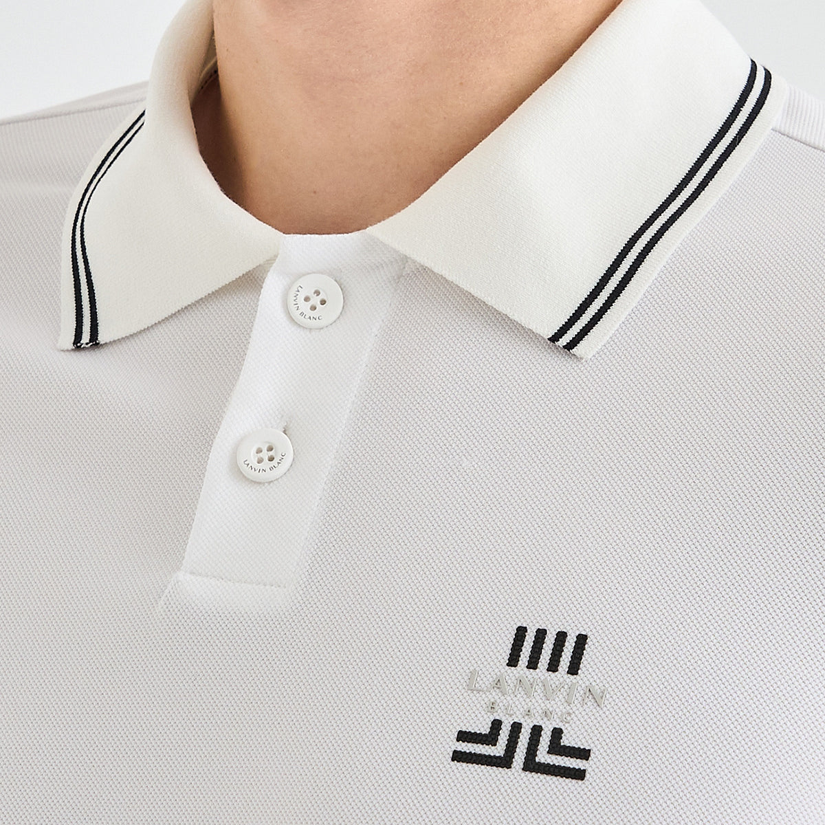 Close-up of a white polo shirt worn by a model, featuring a double-striped collar, white buttons, and a black embroidered 'LANVIN BLANC' logo on the chest.