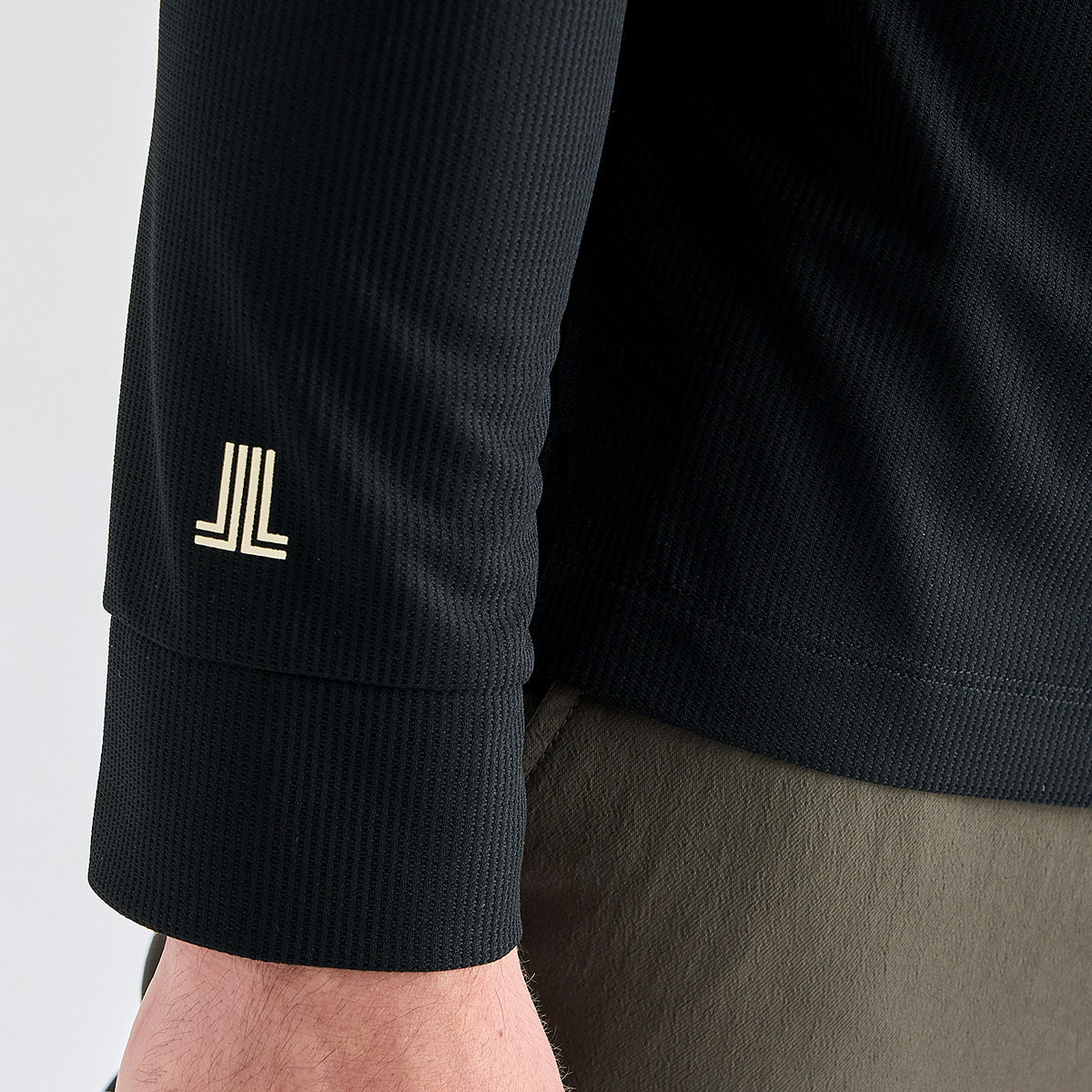 Close-up of a black ribbed long-sleeve athletic shirt worn by a model, showing a gold stylized 'JL' logo on the left sleeve cuff.