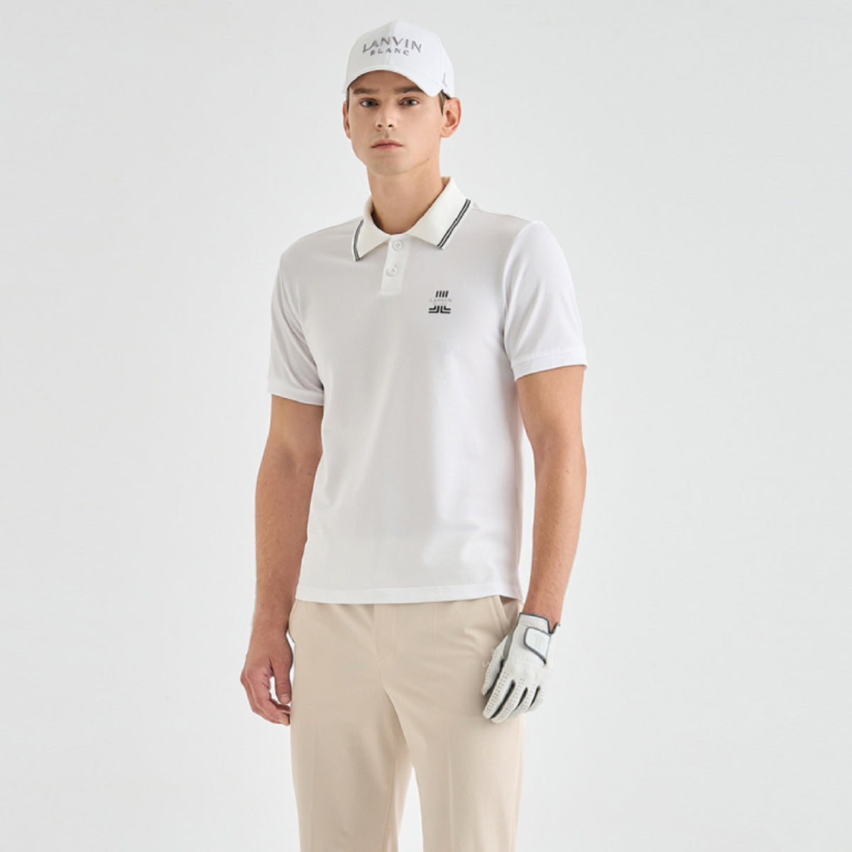 A man modeling a white short-sleeve polo shirt with a dark gray striped collar, paired with beige trousers, a white cap, and a white golf glove against a light gray studio background.