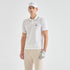 A man modeling a white short-sleeve polo shirt with a dark gray striped collar, paired with beige trousers, a white cap, and a white golf glove against a light gray studio background.
