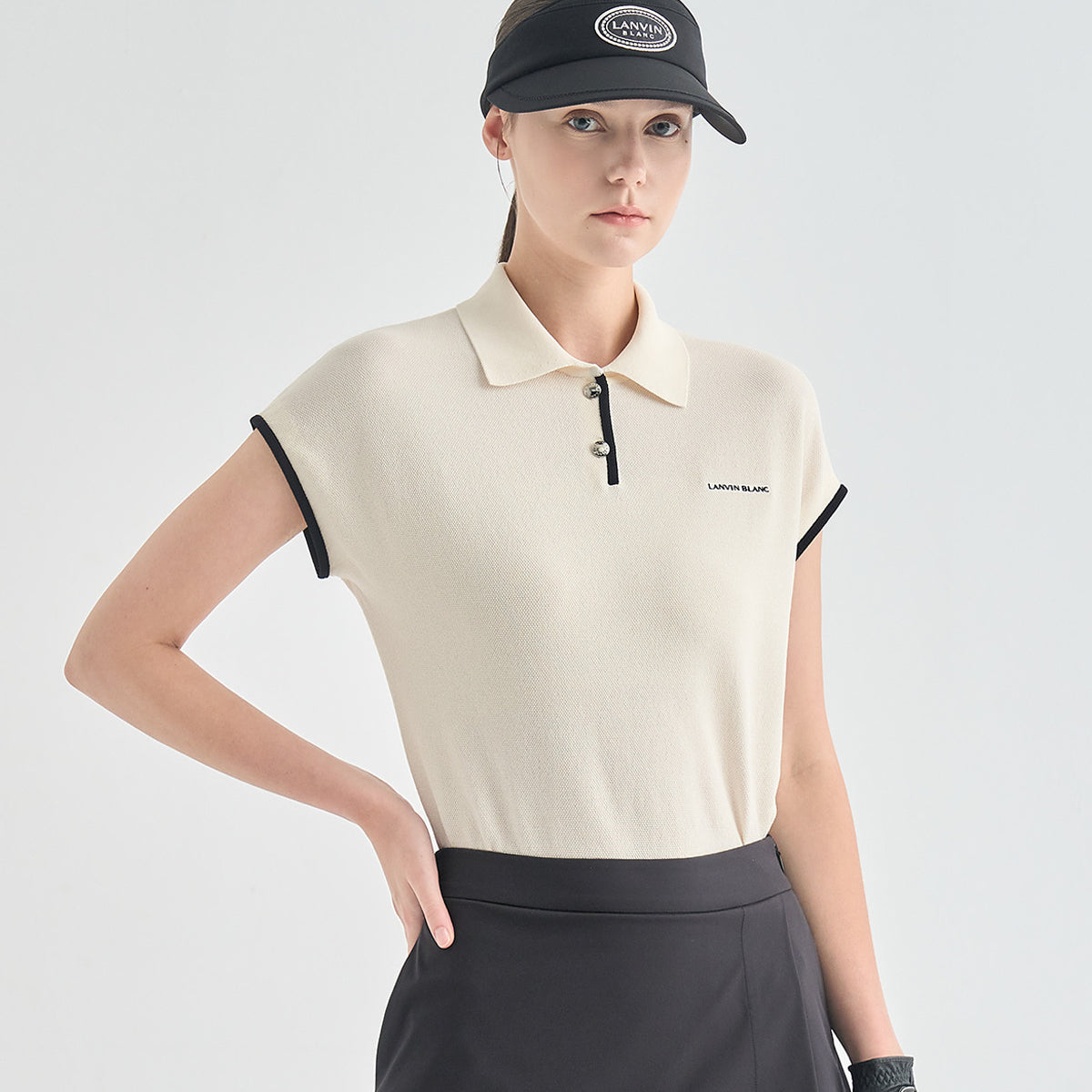 A model wearing a short-sleeve polo shirt in cream with black trim on the collar and sleeve cuffs, paired with a black skirt and a black cap. The brand name 'LANVIN BLANC' is visible on the chest of the polo shirt.