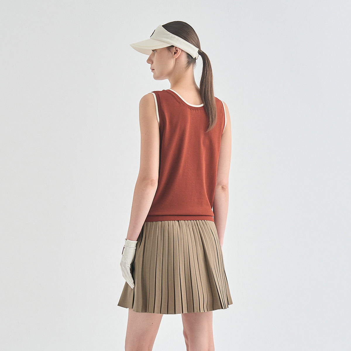 A woman shown from the back wearing a sleeveless rust-orange knit top, a pleated beige tennis skirt, a white visor, and a white glove, set against a plain light gray studio background.