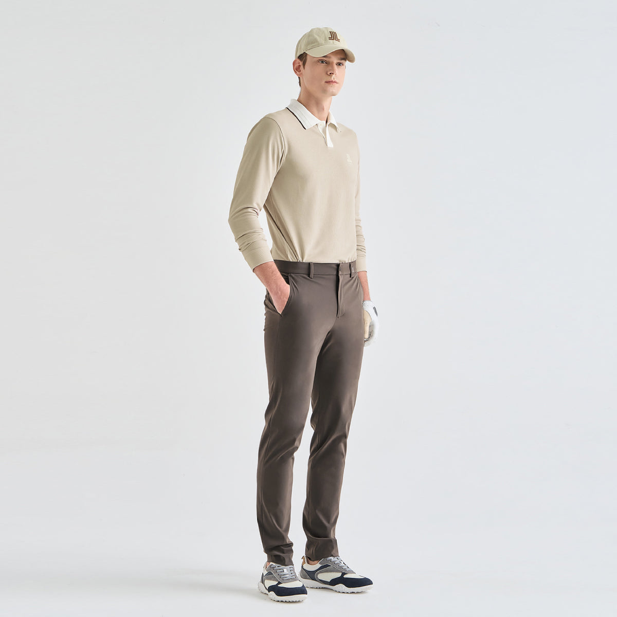 A man modeling a beige long-sleeve polo shirt, brown golf pants, a beige cap, and white golf shoes against a clean white studio background.