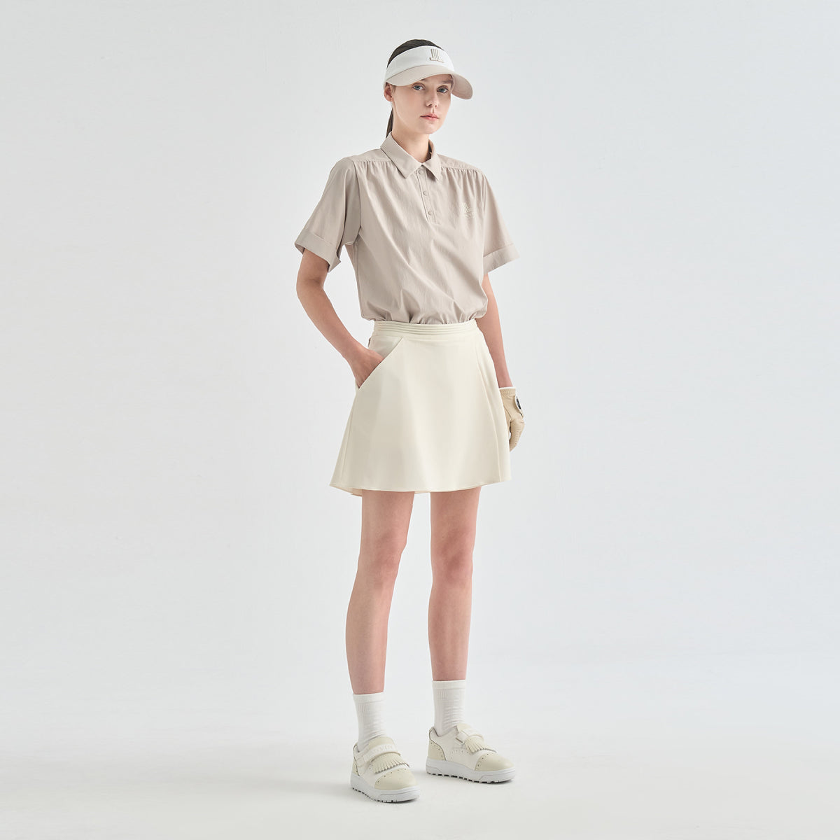 A model stands against a light gray studio background wearing a beige short-sleeve collared shirt, an off-white pleated mini skirt, a beige visor, white socks, and off-white sneakers.