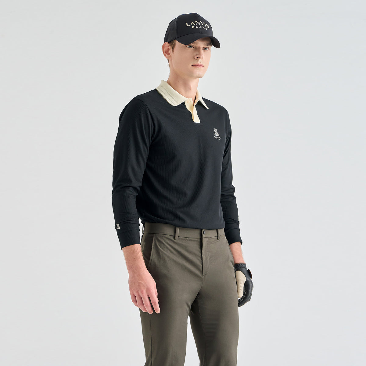 A male model wearing a black long-sleeve golf polo with a cream collar, olive green pants, a black cap with 'LANVIN BLANC' text, and a black glove, photographed against a light gray studio background.