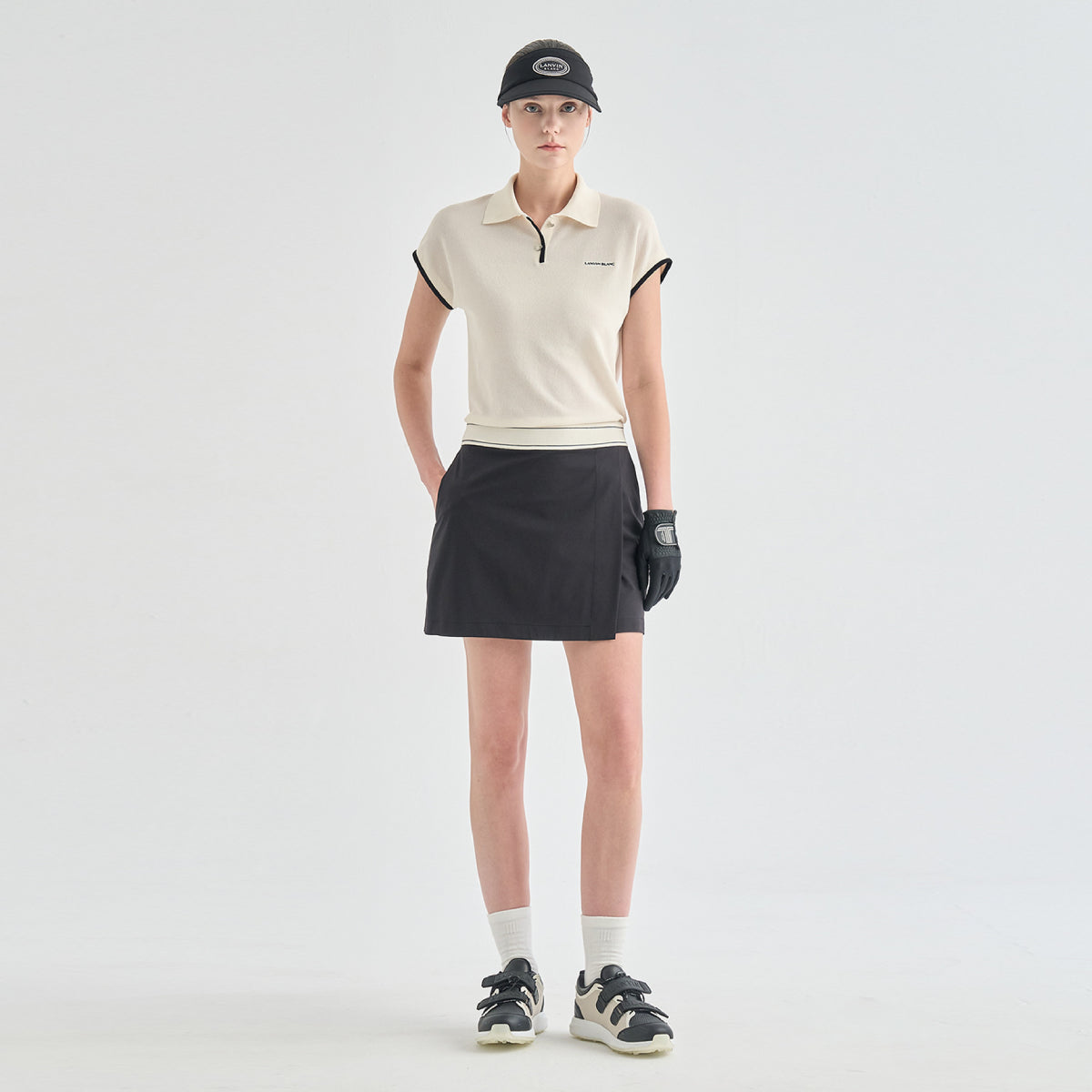 A model stands against a light gray studio background wearing a cream short-sleeve polo shirt with black trim, a black skirt, a black cap, a black glove, white socks, and black-and-white sneakers.