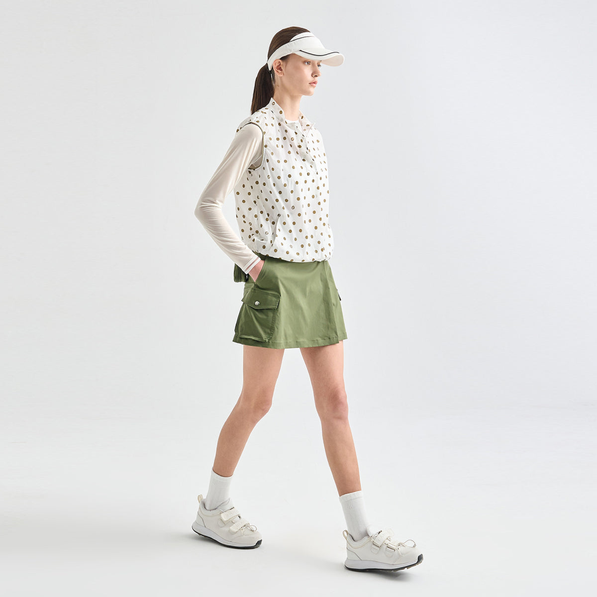 A woman models a sleeveless white button-up shirt with small brown polka dots, worn over a fitted long-sleeve white athletic base layer, paired with an olive green skort and white sneakers, against a light gray studio background.