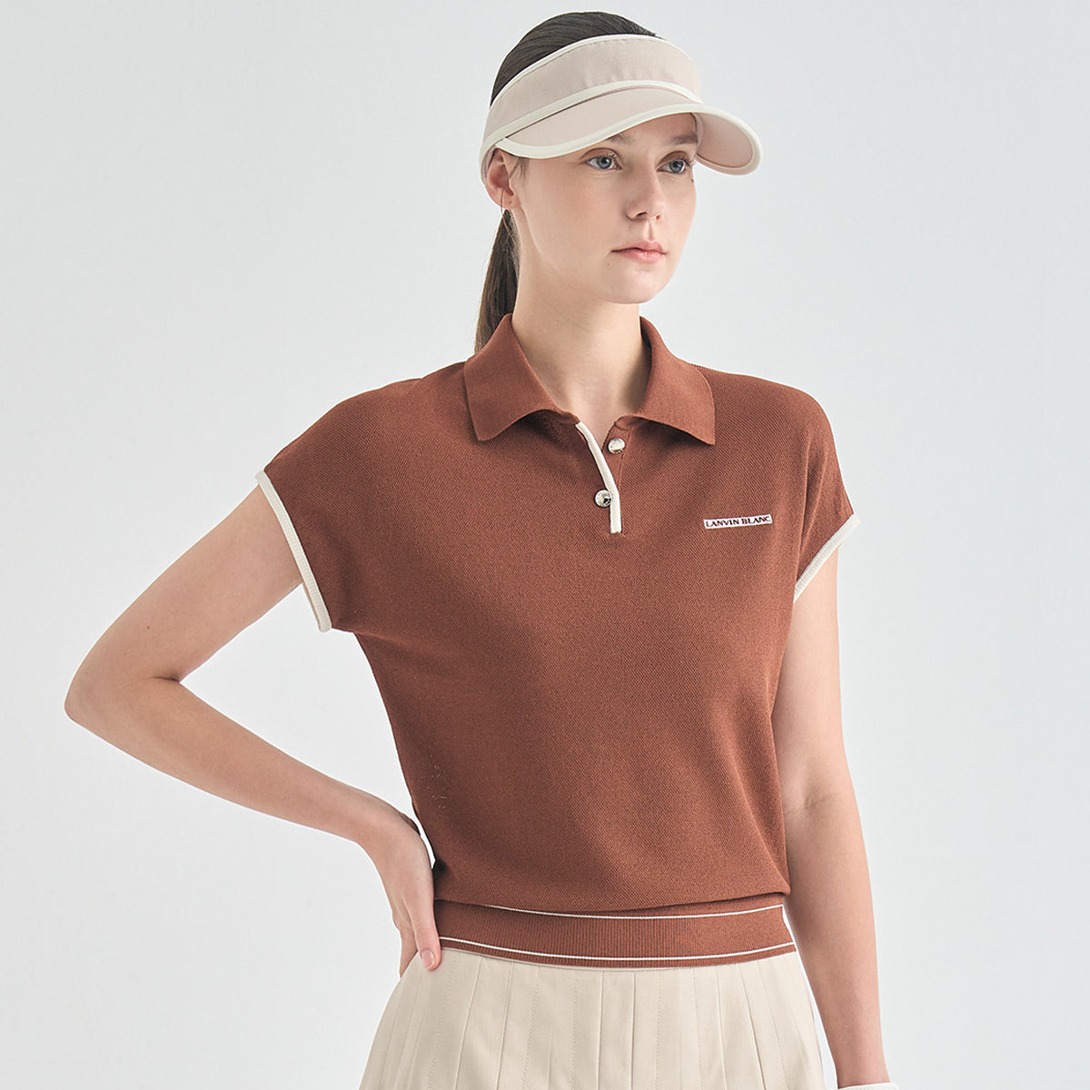 A woman wearing a short-sleeve knit polo shirt in a warm rust brown color, styled with a beige visor and a cream pleated skirt against a light gray studio background.