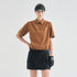 A model wearing a brown short-sleeve collared polo shirt with a small white logo on the left chest, paired with a black skirt, black visor, and black glove, against a plain light gray studio background.