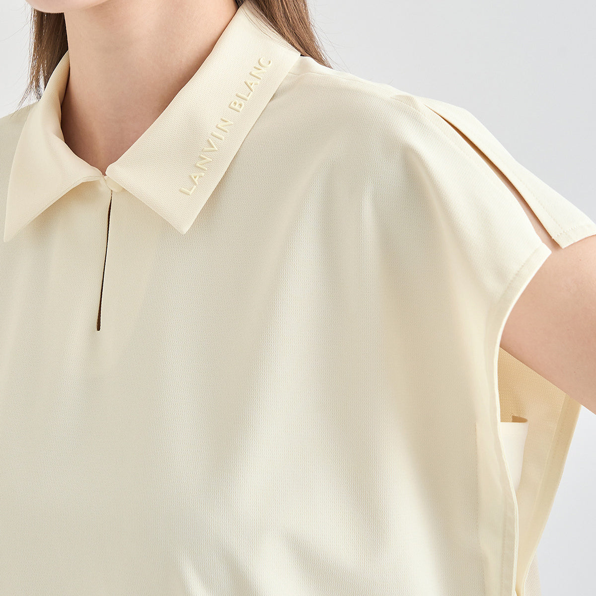 Close-up of a model wearing a pale cream short-sleeve collared top with a subtle V-neckline and 'LANVIN BLANC' text embroidered on the collar, against a light gray background.