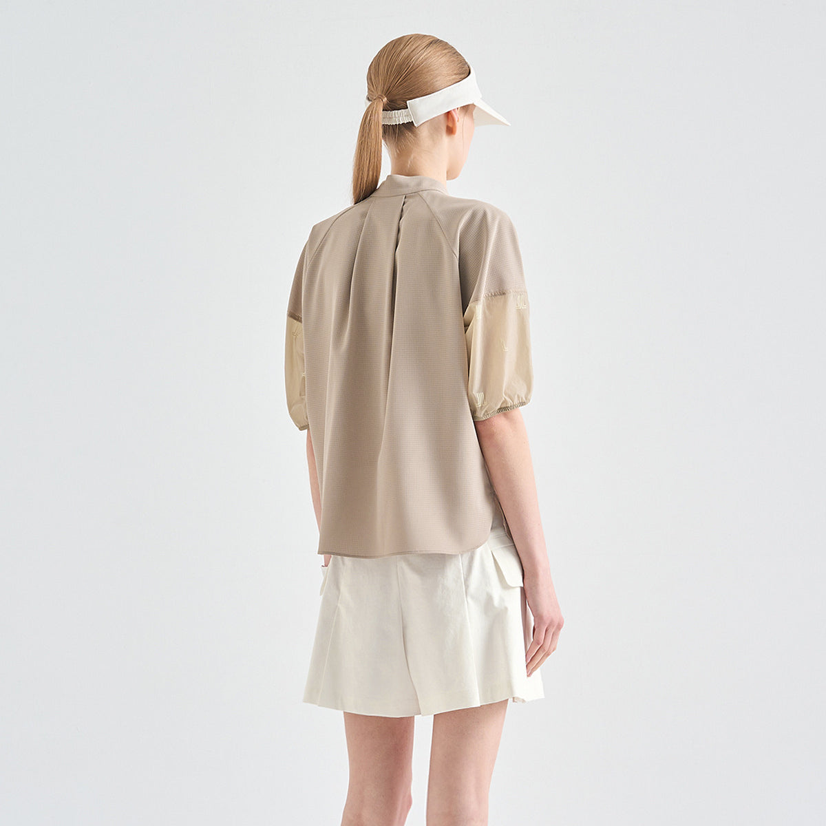 A woman shown from the back wearing a beige short-sleeve blouse with contrasting beige puff sleeves and white shorts, against a light gray studio background.