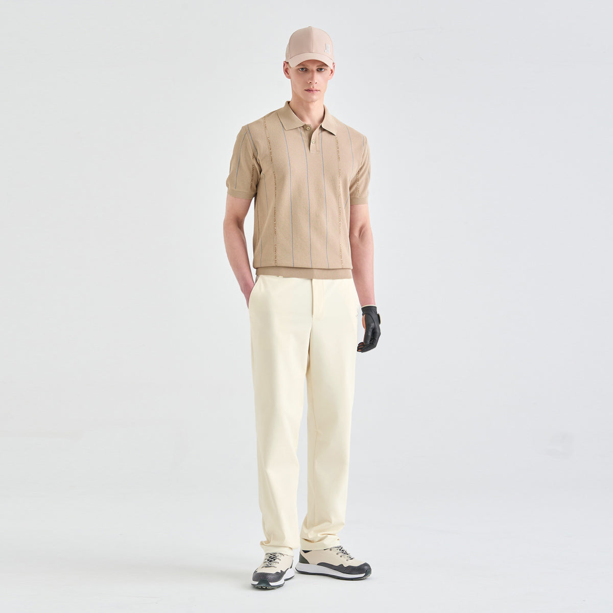 A male model stands against a seamless white studio background wearing a beige short-sleeve collared knit top, cream trousers, a beige cap, and a black glove.