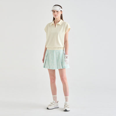A woman modeling a light yellow sleeveless polo shirt and a light green pleated tennis skirt, paired with a white visor, white glove, white socks, and white sneakers, against a plain light gray studio background.
